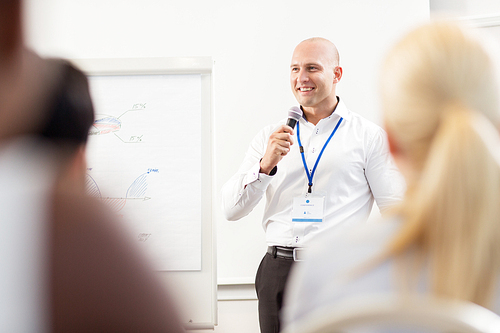 business, education and strategy concept - smiling businessman with microphone and charts on whiteboard talking to group of people at conference presentation