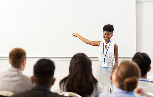 business, education and people concept - smiling african american businesswoman or teacher with remote and group of students at conference presentation or lecture