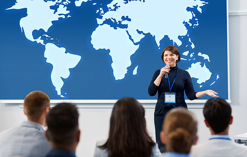 business, education and people concept - smiling businesswoman or lecturer with microphone and world map on projection screen talking to group of students at conference presentation or lecture