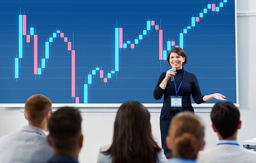 business, economy and people concept - smiling businesswoman or financier with forex chart on projection screen microphone talking to group of students at conference presentation or lecture