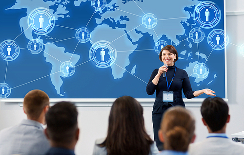 business, global network and people concept - smiling businesswoman or lecturer with world map on projection screen and microphone talking to group of students at conference presentation or lecture