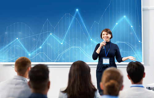 business, statistics and people concept - smiling businesswoman or lecturer with microphone and diagram chart on projection screen talking to group of students at conference presentation or lecture