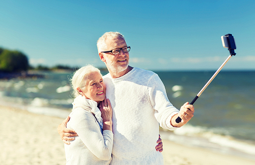 family, age, travel, technology and people concept - happy senior couple with smartphone selfie stick photographing and hugging on summer beach