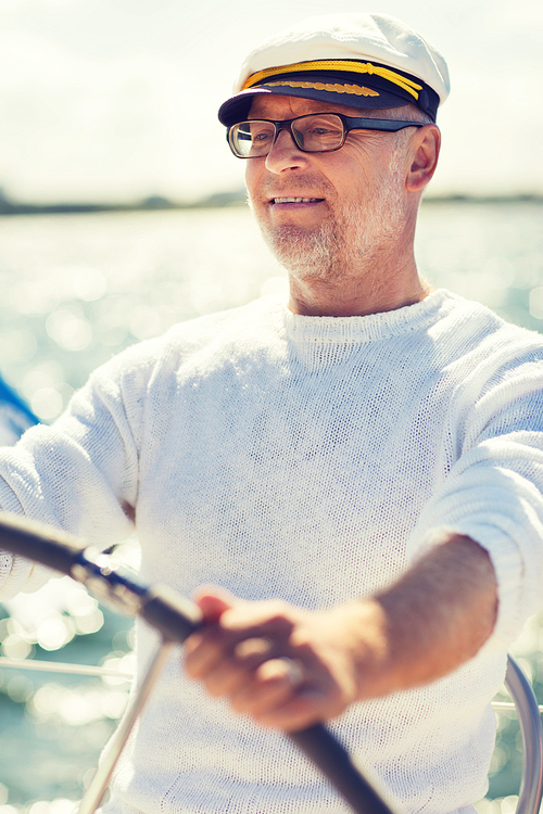 sailing, age, tourism, travel and people concept - happy senior man in captain hat on steering wheel and navigating sail boat or yacht floating in sea