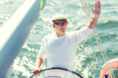 sailing, age, tourism, travel and people concept - happy senior man in captain hat on steering wheel and navigating sail boat or yacht floating in sea