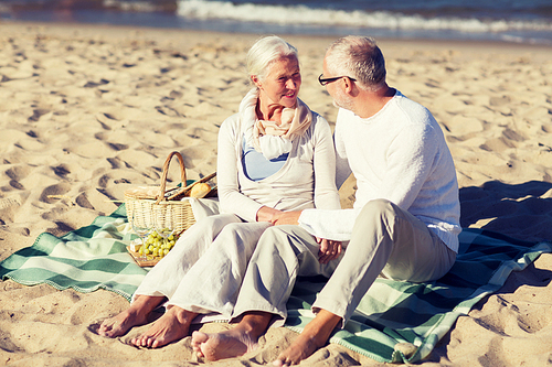 family, age, holidays, leisure and people concept - happy senior couple with picnic basket sitting on blanket on summer beach