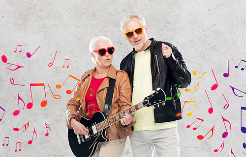 music, age and people concept - happy senior couple in sunglasses with electric guitar over gray concrete background and musical notes