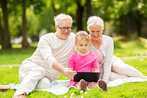 family, generation and technology concept - happy smiling grandmother, grandfather and little granddaughter with tablet pc computer sitting on blanket at park