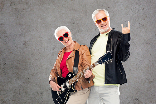 music, age and people concept - happy senior couple in sunglasses with electric guitar showing rock hand sign over gray concrete background