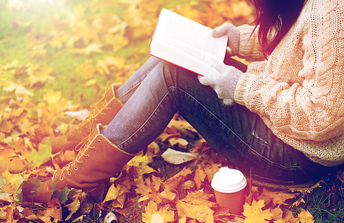 season, literature, education and people concept - close up of young woman reading book and drinking coffee from paper cup in autumn park