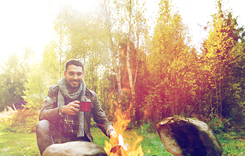 camping, travel, tourism, hike and people concept - happy man drinking hot tea near fire at camp in woods