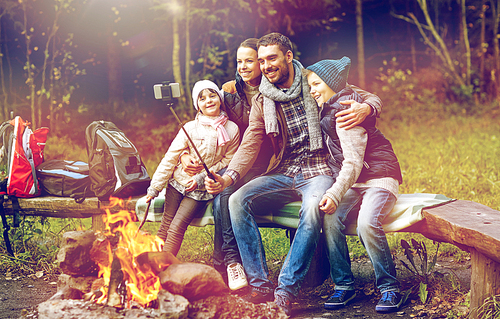hike, travel, tourism and people concept - happy family sitting on bench and taking picture with smartphone on selfie stick at campfire in woods