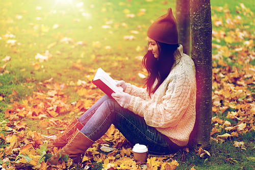 season, literature, education and people concept - young woman reading book and drinking coffee from paper cup in autumn park