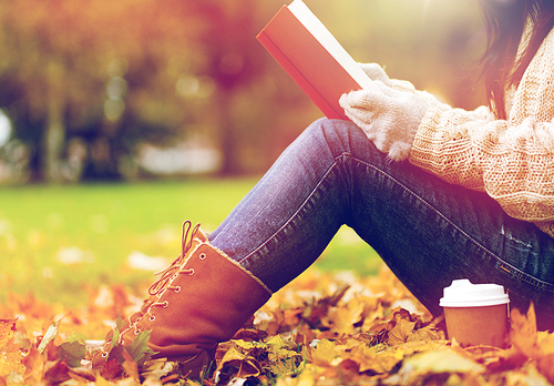 season, literature, education and people concept - close up of young woman reading book and drinking coffee from paper cup in autumn park