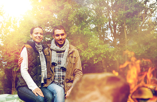 travel, tourism and hike concept - happy couple sitting on bench and warming near campfire at camp in woods