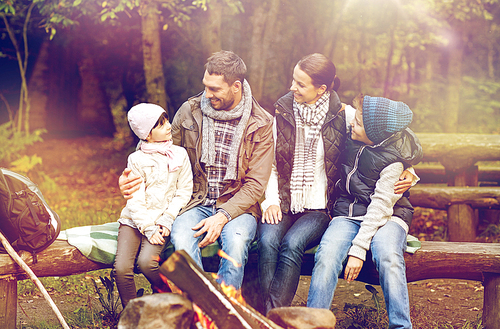 hike, travel, tourism and people concept - happy family sitting on bench and talking at camp near campfire in woods