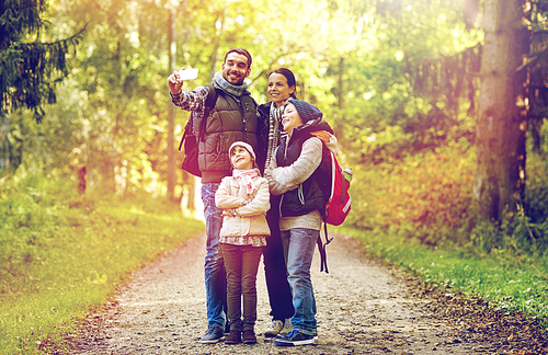 travel, tourism, hike, technology and people concept - happy family with backpacks taking selfie by smartphone in woods