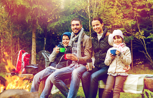 travel, tourism, hike and people concept - happy family sitting on bench and drinking hot tea from cups at camp fire in woods