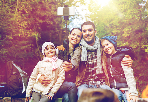 hike, travel, tourism and people concept - happy family sitting on bench and taking picture with smartphone on selfie stick at campfire in woods