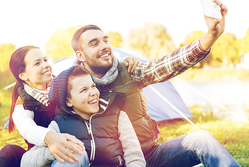 hike, travel and technology people concept - happy family with smartphone taking selfie at campsite