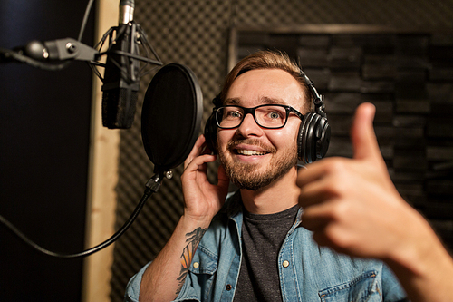 music, show business, people and voice concept - male singer with headphones and microphone singing song at sound recording studio and showing thumbs up