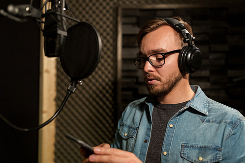 music, show business, people and voice concept - male singer with headphones, microphone and smartphone at sound recording studio