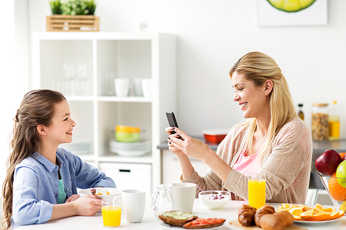family, technology and people concept - happy girl and mother with smartphone having breakfast at home