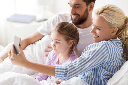 people, family and technology concept - happy mother, father and little girl taking selfie with smartphone in bed at home