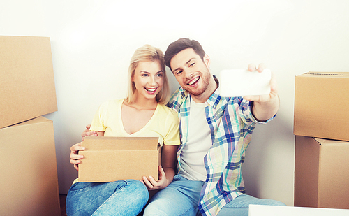 home, people, repair and real estate concept - smiling couple with big cardboard boxes moving to new place and taking smartphone selfie