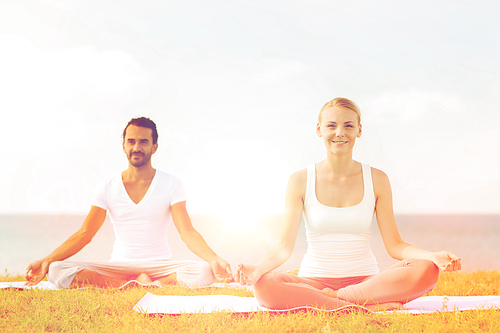 fitness, sport, friendship and lifestyle concept - smiling couple making yoga exercises sitting on mats outdoors