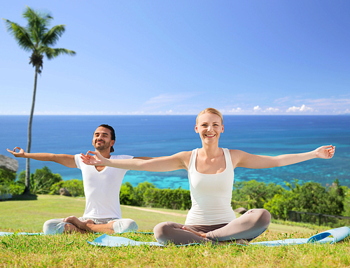 fitness, sport, yoga and people concept - happy couple meditating in lotus pose outdoors over exotic natural background with palm tree and ocean