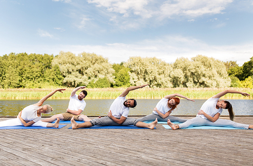fitness, sport, and healthy lifestyle concept - group of people making yoga exercises outdoors on river or lake berth