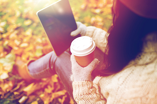 season, technology and people concept - young woman with tablet pc and coffee cup sitting on grass in autumn park