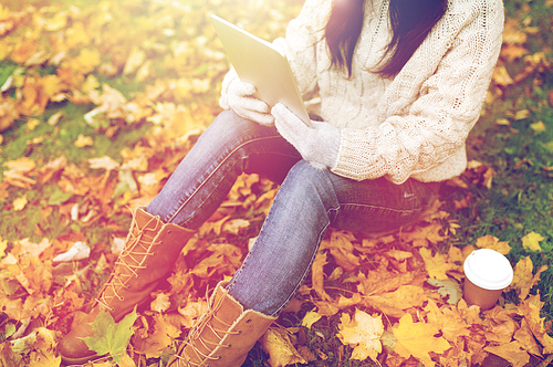 season, technology and people concept - young woman with tablet pc and coffee cup sitting on grass in autumn park