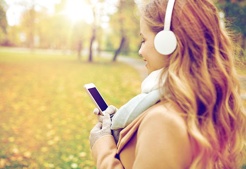 season, technology and people concept - close up of beautiful happy young woman with headphones listening to music on smartphone walking in autumn park