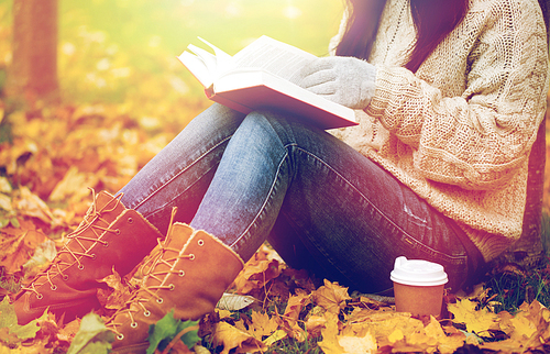 season, literature, education and people concept - close up of young woman reading book and  coffee from paper cup in autumn park