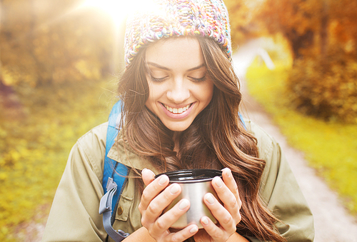 adventure, travel, tourism, hike and people concept - smiling young woman with cup and backpack in autumn forest