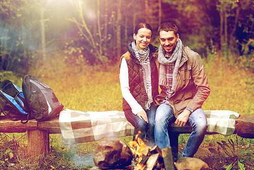 travel, tourism, hike and people concept - happy couple sitting on bench and warming near campfire at camp in woods