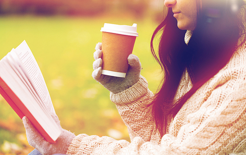 season, literature, education and people concept - close up of young woman reading book and drinking coffee from paper cup in autumn park