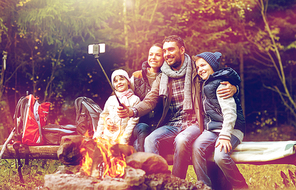 travel, tourism and hike concept - happy family sitting on bench and taking picture by smartphone on selfie stick at campfire in woods
