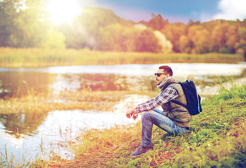 travel, tourism, hike and adventure concept - man in sunglasses with backpack resting on river bank