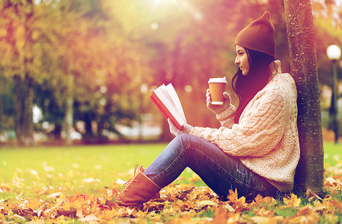 season, literature, education and people concept - young woman reading book and drinking coffee from paper cup in autumn park