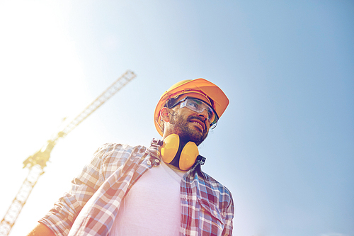 building and people concept - smiling builder with hardhat and headphones over blue sky and construction crane