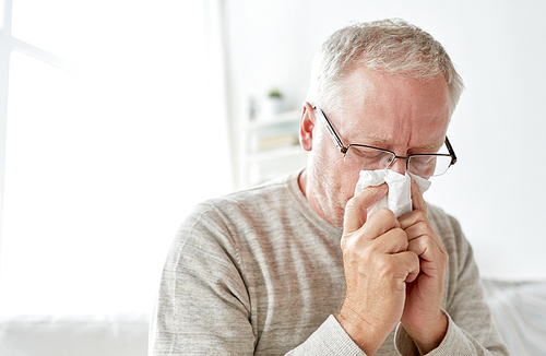 healthcare, flu, hygiene and people concept - sick senior man with paper wipe blowing his nose at home