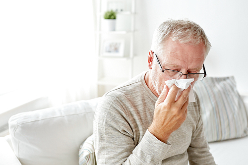 healthcare, flu, hygiene and people concept - sick senior man with paper wipe blowing his nose at home