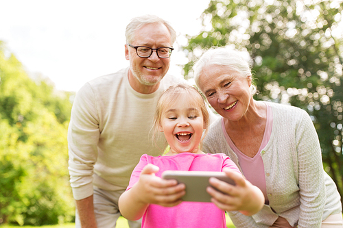 family, generation and people concept - happy smiling grandmother, grandfather and little granddaughter taking selfie by smartphone at park