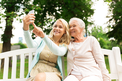 family, technology and people concept - happy smiling young daughter and senior mother with smartphone sitting on park bench and taking selfie