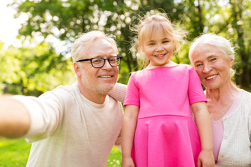 family, generation and people concept - happy smiling grandmother, grandfather and little granddaughter taking selfie at park