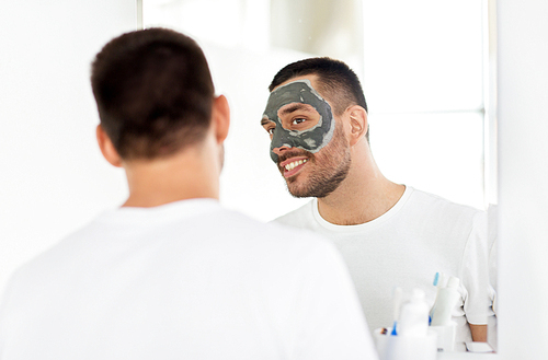 grooming, skin care and people concept - young man with clay mask on face and looking to mirror at home bathroom