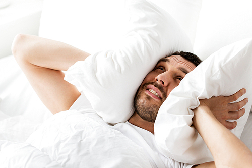 people, bedtime and rest concept - man lying in bed with pillow suffering from noise at home